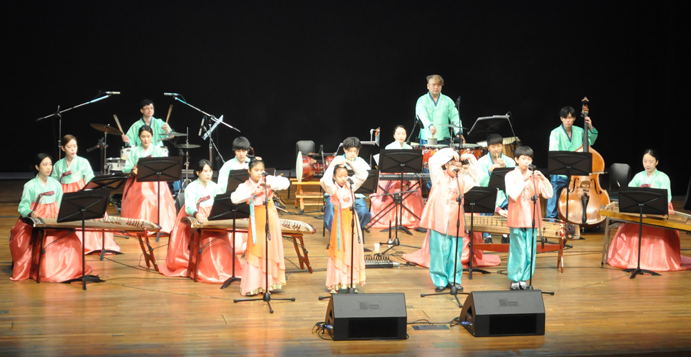 한복을 곱게 차려입은 4명의 아이들이 무대에서 노래하고 그 뒤로 11명의 연주자가 전통악기를 연주하고 있다
