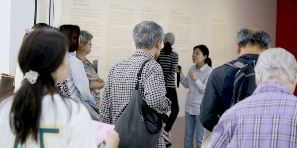 전시장을 안내하고 있는 담당 학예사를 8명의 관람객이 바라보며 전시장을 둘러보고 있다.