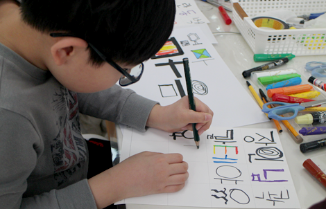 초등학생 남자 어린이가 왼손으로 색연필을 잡고 종이 위에 한글 글자를 쓰고 있다.