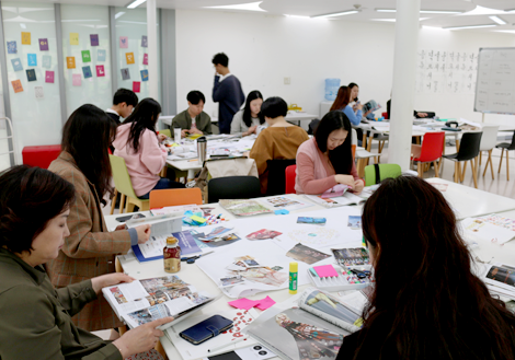 한글 상표 공방 수업 중 자신의 작품을 만드는 학생들의 모습. 테이블 위로 디자인 관련 서류들이 어지러이 늘어져 있고, 12명의 학생이 자신만의 작업에 몰두해 있다.