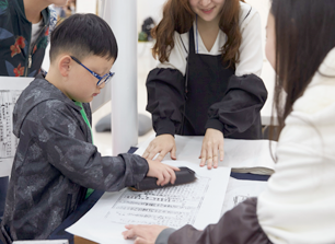 한 어린이가 목판인쇄 체험에 나서 종이를 문지르고 있다. 양 옆에서 두 명의 진행자가 아이를 돕고 있다.