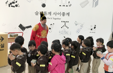 세종대왕으로 분장한 연기자가 16명의 어린이들을 뒷짐을 진 채 흐뭇하게 바라보고 있다.