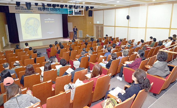 50여 명이 강당의 자리에 앉아 무대 위에서 ‘해부학 이야기’를 강연하는 한 남성의 강연을 경청하고 있다.