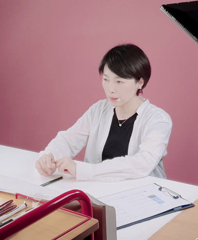 단발 머리의 한 여성이 분홍색 배경의 스튜디오에 앉아 촬영을 하고 있다. 흰색 책상 위에는 촬영에 참고할 서류와 펜이 놓여져 있다. 