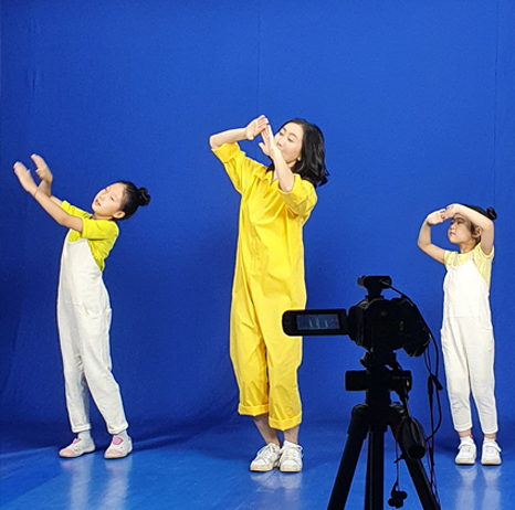 블루스크린이 설치된 촬영장에서 촬영이 진행되고 있다. 노란색 옷을 입은 여성과 양 옆으로 두 명의 여자아이가 하늘을 보며 손을 모으는 포즈를 취하고 있다.