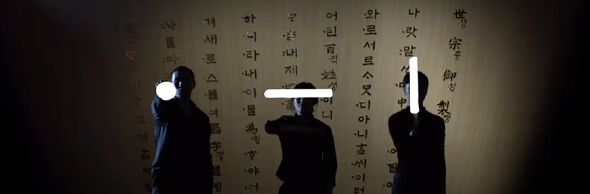 세 사람이 나란히 서 있다. 그들의 얼굴 위로는 모음 ‘ㆍ’, ‘ㅡ’, ‘ㅣ’가 하얀 빛처럼 떠 있다.