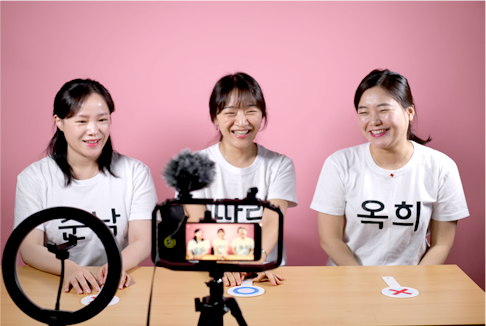 흰 티셔츠를 입은 세명의 여성이 나란히 앉아 한 곳을 바라보며 웃고 있다. 여성의 앞에는 마이크와 촬영용 휴대폰이 삼각대 위로 세팅되어 있다.
