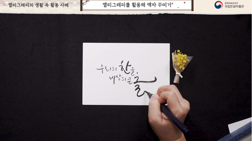 캘리그래피를 활용해 액자 꾸미기 페이지. 하얀 종이 위에 ‘우리의 한글, 세상의 큰글’이라 캘리그래피를 적는 모습. 