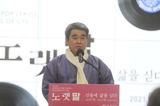 박재혁 부산박물관 학예연구실장이 개회사에 나선 모습. ‘노랫말 - 선율에 삶을 싣다’라고 적힌 단상 위의 마이크에 입을 가져다대고 정면을 바라보며 말하고 있다. 보랏빛 한복 위에 하얀 목도리를 착용했다. 