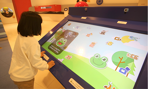 한글놀이터 체험 디지털기기 앞에 서서 한 여자 어린이가 개구리 그림을 터치해보고 있다.