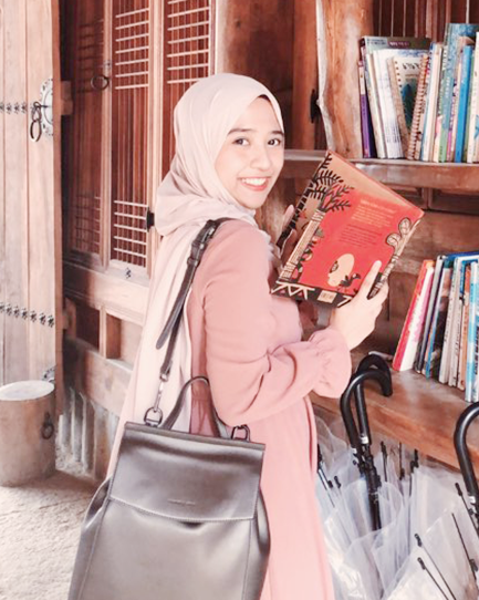 히잡을 쓰고 오른쪽 어깨에는 검은색 숄더백을 맨 체 손에는 책을 펼쳐 들고 카메라를 바라보며 웃는 샤이라.