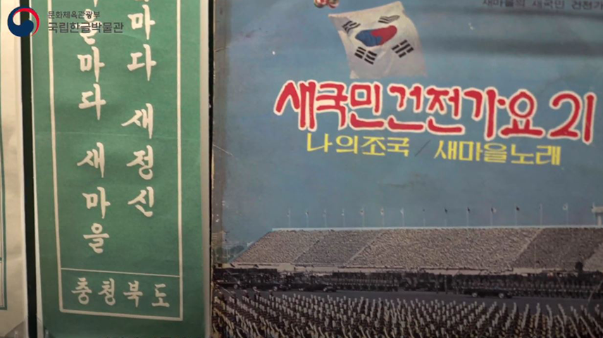 1960년대 새마을운동과 관련된 표어. 왼편에는 녹색 바탕에 세로쓰기로 표어가 적혀 있다. 윗부분이 잘려 ‘마다 새정신’, ‘을마다 새마을-충청북도’만 보인다. 오른쪽 포스터에는 태극기가 휘날리는 경기장을 바탕으로 ‘새국민건전가요21’, ‘나의조국/새마을노래’가 적혀 있다.