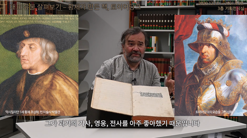 “기획특별전 ‘문자 혁명-한국과 독일의 문자 이야기’_대표 유물 살펴보기Ⅱ” 영상 캡처. 길게 기른 머릴 뒤로 묶고 턱수염을 기른 외국인 남성이 커다란 책을 앞에 두고 설명하고 있다. 뒤로는 빼곡하게 책이 꽂힌 책꽂이가 있다. 남성의 왼쪽엔 막시밀리안 1세 황제 초상화 그림이 합성되어 있다. 초상화 속 인물은 귀를 덮는 하얀 단발머리에 검은색 챙이 넓은 모자를 썼다. 검은 옷 위에 두툼해 보이는 갈색 털코트를 걸치고 있다. 오른쪽에는 토이어당크의 모습을 그린 삽화가 합성되어 있다. 토이어당크의 뒤로 푸른 하늘과 빨간 커튼이 배경으로 그려져 있다. 토이어당크는 왼편을 바라보고 있으며, 금색으로 화려하게 무늬가 새겨진 은색 갑옷을 입고 있다. 머리에도 비슷한 무늬가 새겨진 투구를 쓰고 있으며 투구 위에는 파란색, 흰색, 빨간색이 섞인 장식물과 왕관처럼 생긴 장식물이 올려져 있다. 영상 아래 자막에는 ‘그가 과거의 기사, 영웅, 전사를 아주 좋아했기 때문입니다’라고 적혀있다.