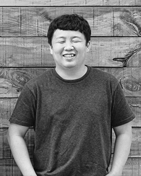 흑백사진으로 반팔 티셔츠를 입은 김용관 작가가 나무 벽면 앞에서 활짝 웃고 있다. 