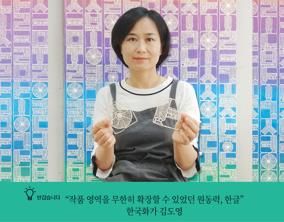 반갑습니다 <“작품 영역을 무한히 확장할 수 있었던 원동력, 한글” 한국화가 김도영> 기사 사진. 직접 만든 한글 한옥 작품을 손에 든 김도영 작가가 살짝 미소지은 채 카메라를 응시하고 있다. 뒤로는 그의 작품이 배경으로 활용됐다. 보라색, 하늘색, 청록색으로 차례대로 칠해진 배경에 한글 자음이 빼곡하게 새겨진 작품이다.