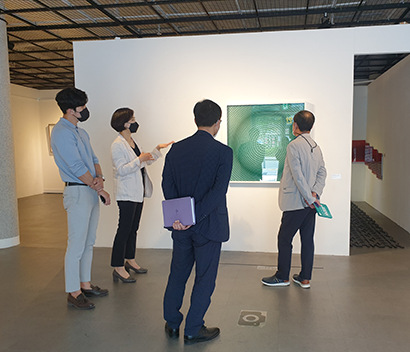 전시장 벽면에 작품이 걸려 있고, 그 앞에 네 명의 사람들이 서 있다. 하얀색 자켓과 검은색 바지를 입은 김도영 작가가 작품을 설명하고 있으며, 나머지 세 명의 사람들은 그 설명을 듣고 있다. 