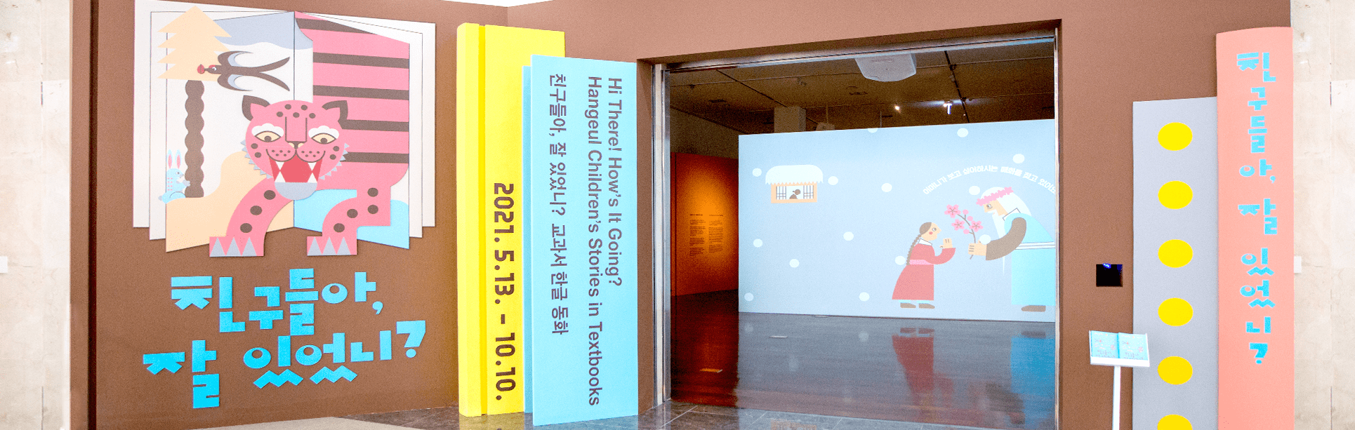 ‘친구들아, 잘 있었니? 교과서 한글 동화’ 전시장 입구 전경. 책장에 책이 꽂혀진 것처럼 꾸며져 있다. 책모양 패널에는 ‘친구들아, 잘 있었니?’, ‘HI, There! How’s It Going? Hangeul Children’s Stories in Testbooks’, ‘2021.5.13. - 10.10.’이 적혀있다. 입구 왼편에는 익살스럽게 생긴 호랑이가 책 속에서 막 뛰어나올 것 같은 그림과 함께 전시 제목 ‘친구들아, 잘 있었니?’가 적혀있다. 입구 너머로 전시장이 보인다.