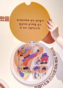 전시장에서 만나 볼 수 있는 체험형 전시물. 벽면에 설치된 동그란 판 위에 노란색 덮개가 덮여있는 구조물이다. 덮개를 열면 덮개에 적힌 전래동화 문구를 읽을 수 있다. 동그라미 속에는 문구 내용과 관련된 그림이 그려져 있다. 덮개에 적힌 문구는 ‘무시무시하게 생긴 장사들이 몽둥이와 올가미를 들고 한 무리 나왔다는구먼’이며, 동그라미 속에는 놀부를 혼내는 장사 그림과 흥부와 놀부가 서로 웃으며 껴안고 있는 그림이 그려져 있다. 동그라미 아래에는 모양을 따라 문구가 적혀있는데 ‘놀부네 첫째 박이 갈라지고 뭐가 나왔’까지만 보인다.