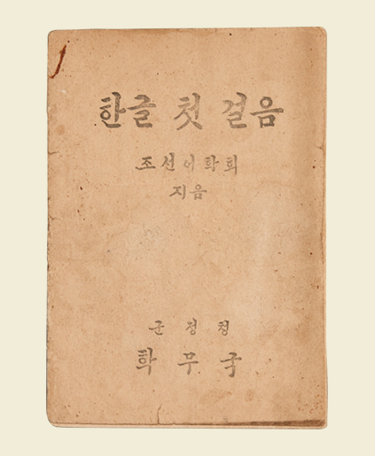 ‘한글 첫 걸음’ 표지. 붉은 갈색으로 빛바랜 표지에 ‘한글 첫 걸음’이 적혀있다. 그 아래에는 ‘조선어학회 지음’이 적혀있으며 표지 하단에는 ‘군정청/ 학무국’이라고 적혀있다. 글씨체는 붓글씨체와 비슷하다.