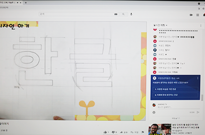 실시간으로 송출되고 있는 원격 교육 화면. 영상에서 연필로 스케치 된 ‘한글’ 글자가 보인다. 
영상 옆에는 실시간 채팅창으로 수업에 참여하는 아이들의 채팅이 적혀있다. 글자에 대한 느낌이 어떤지 투표하는 실시간 투표창도 채팅창에 함께 올라와 있다.