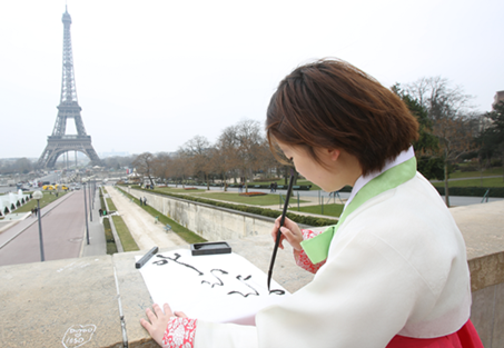 한복을 차려입은 이정화 작가가 길거리에서 붓글씨를 쓰고 있다. 저 멀리 파리의 에펠탑이 보인다.
