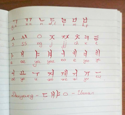 노트 위에 서툰 글씨로 한글 자음과 모음이 나란히 적혀있다. 한글 아래에는 영어로 자음의 발음이 적혀있다. 글씨는 빨간색 펜으로 적혔다. 하단에는 ‘Daeyong’, ‘ㄷㅐㅑㅇ’, ‘Umman’ 등이 적혀있다.