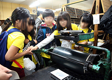 어린이들이 활판공방에서 활판 인쇄와 관련된 기계를 직접 만지며 체험하고 있다. 대여섯 명의 어린이가 노란색 유치원 옷을 입고 기계 앞에 함께 모여 서 있다.