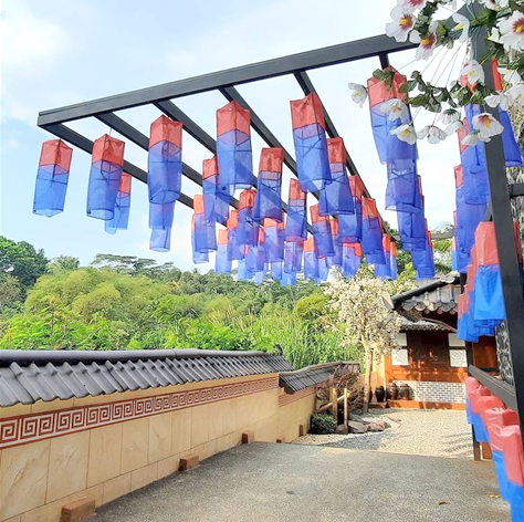 낮은 한옥 담장과 한국식 마당이 있고, 마당에는 빨간색과 파란색이 섞인 청사초롱이 빼곡하게 설치되어 있다. 마당 너머로는 한옥과 울창한 산이 보인다.