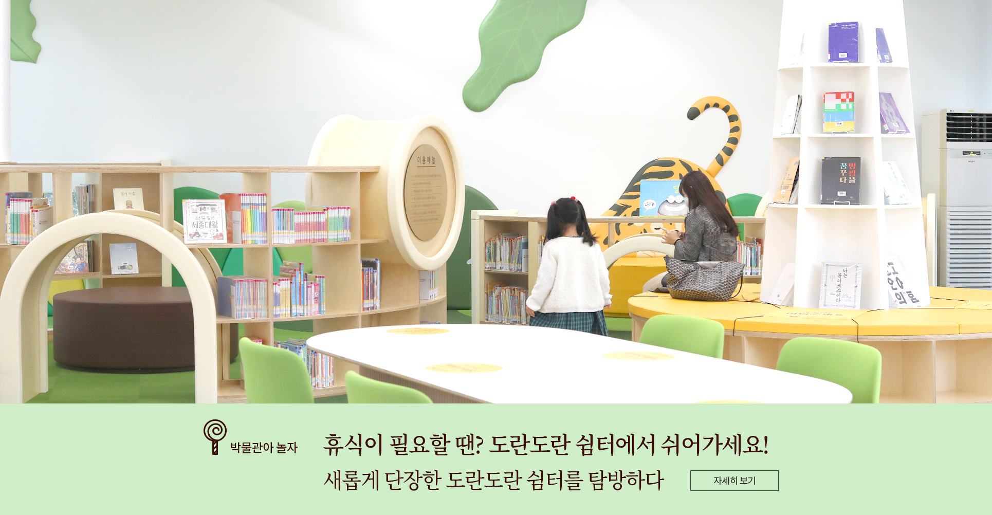 박물관아 놀자 코너 기사 사진. 도란도란 쉼터 실내 사진. 앞에는 탁자와 의자가 놓여있고, 그 너머로 아이들이 즐길 수 있는 서가 공간이 보인다. 책이 잔뜩 꽂힌 책꽂이가 서가를 두르고 있다. 왼편에는 높은 기둥을 중심으로 설치된 노란색 소파가 있고 한 여성 관람객이 앉아 핸드폰을 하고 있다. 기둥에는 책이 표지가 보이도록 전시되어 있다. 여성의 왼쪽에는 머리를 양 갈래로 묶고 하얀색 가디건을 입은 여자아이가 서 있다. 아이와 관람객 모두 뒷모습만 보인다. 사진 하단에는 박물관아 놀자 기사 제목인 ‘휴식이 필요할 땐? 도란도란 쉼터에서 쉬어가세요! 새롭게 단장한 도란도란 쉼터를 탐방하다’가 적혀있다. 