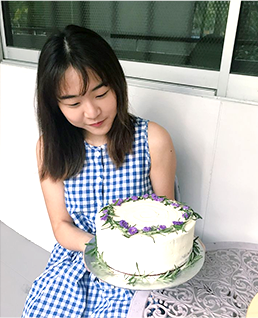 파란색 체크무늬의 원피스를 입은 데프니가 자신이 만든 케이크를 들고 있다. 은색 쟁반 위에는 흰색 크림, 보라색 꽃으로 장식된 케이크가 올려져 있다.
