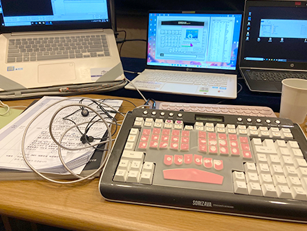 책상 위에 노트북 세 대와 속기 타자기가 놓여있다. 노트북 화면에 속기 프로그램이 열려 있다. 속기 타자기 왼쪽에는 이어폰과 행사 자료가 놓여있다.