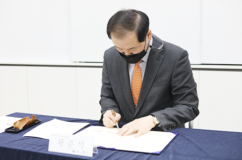황준석 국립한글박물관장이 업무협약서에 서명하고 있다. 그는 회색 재킷과 주황색 넥타이, 검은색 마스크를 착용하고 있다.