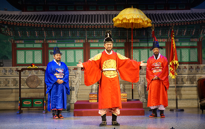 궁궐을 배경으로 한 무대 위에 세 명의 공연자들이 서 있다. 가운데 공연자는 붉은색 곤룡포를 입고 있다. 양옆의 공연자들은 각자 파란색, 빨간색의 조선시대 관료 복장을 하고 있다.