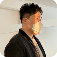 김동관 디자이너의 측면 사진이다. 그는 검은색 재킷을 입고 하얀색 마스크를 착용했다. 그는 오른쪽 어딘가를 응시하고 있으며, 그의 얼굴에 틈새로 비친 햇볕이 드리워져 있다.