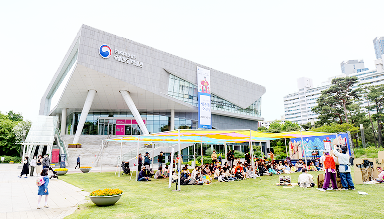 국립한글박물관 외관 전경이다. 한글박물관 건물에는 ‘세종대왕 오신 날’ 기념 천막이 크게 걸려있다. 건물 앞 잔디밭에는 커다란 천막이 설치되어 있고, 천막 아래 행사에 참여하는 관람객들이 모여 앉아있다. 관람객 앞에선 한복을 입은 배우들이 전통악기를 곁에 둔 채 연기를 펼치고 있다.