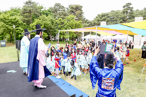 잔디밭에 설치된 무대에 조선 시대 선비 복장과 관료 복장을 한 배우들이 두루마리를 든 채 서 있다. 잔디밭에는 천막이 설치되어 있고, 유생 복장을 한 관람객들이 행사에 일어서서 행사에 참여하고 있다.