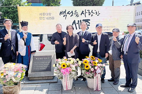 세종대왕 생가터에서 황준석 국립한글박물관장과 국립한글박물관 후원회 이사들이 모여 사진을 찍고 있다. 그들은 대부분 어두운색의 정장을 착용하고 있으며, 한 남성은 조선 시대 선비 복장을 하고 있다. 이들은 ‘세종대왕 탄신 625돌 백성을 사랑한 세종’ 천막을 배경으로, 앞에는 커다란 꽃 화분을 놓은 채, 기념비 옆에 나란히 서 있다. 모두 손을 들어 작은 하트를 만들어 보인다.