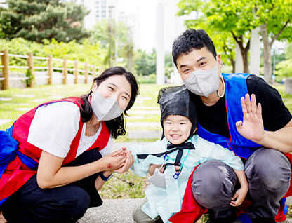 잔디밭에 유생 복장을 한 어린이와 한복 조끼를 입은 부모가 함께 쭈그리고 앉아있다. 어머니는 아이의 손을 꼭 잡고 있으며 아이는 아버지 품에 안겨 기대있다. 아버지는 카메라를 향해 손을 흔들고 있다. 세 가족 모두 환하게 미소짓고 있다.