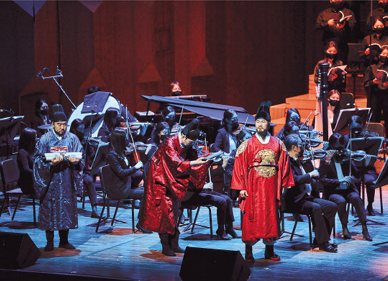 어두운 무대 위로 오케스트라와 연기를 펼치는 배우들이 함께 공연을 하고 있다. 은색 용포를 입은 배우와 각각 붉은색과 푸른색의 관료복을 입은 조선 시대 관료가 연주를 하는 오케스트라 앞에서 열연을 펼치고 있다.