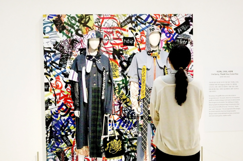 현대적이고 캐주얼한 일상복에 한글디자인이 활용된 작품을 입고 있는 마네킹이 서 있으며, 그 앞에서 한 관람객이 마네킹을 관찰하고 있다. 관람객의 뒷모습만 보인다.