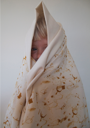 한 외국인 아이가 연한 베이지색 담요를 온몸에 두르고 있다. 담요 사이로 살짝 보이는 아이는 미소를 짓고 있다. 담요에는 갈색의 그림 서체들이 두서없이 새겨져 있다.