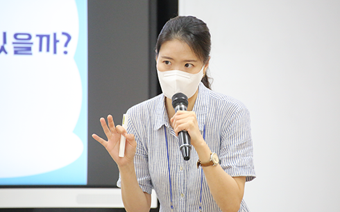 우상미 강사가 한 손에는 펜을, 한 손에는 마이크를 든 채 무언가 말하며 수업을 진행하고 있다. 그녀는 긴 머리를 하나로 묶고 손목에 손목시계를 착용하고 있으며, 하늘색 반팔 원피스를 입고 있다. 그녀는 하얀색 마스크를 착용하고 있다.