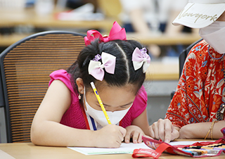 분홍색 원피스를 입은 아이가 연필을 들고 무언가 집중해서 적고 있다. 아이는 마스크를 착용했으며 머리에 세 개의 분홍색 리본을 매달고 있다. 아이의 오른편에서는 아이의 보호자가 함께 앉아 종이 위를 가리키고 있다.