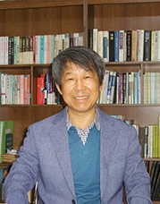 김만태 교수 사진. 책이 가득 꽂힌 책장 앞에서 그가 환하게 미소 짓고 있다. 회색빛 머리의 그는 체크셔츠와 하늘색 스웨터, 푸른색 재킷을 입고 있다.