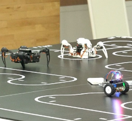 이리저리 하얀 곡선들이 그려져 있는 바닥 위에 로봇 세 대가 놓여있다. 두 대는 다리가 4개 달린 거미처럼 생겼고 한 대는 두 개의 바퀴가 달린 자동차처럼 생겼다.