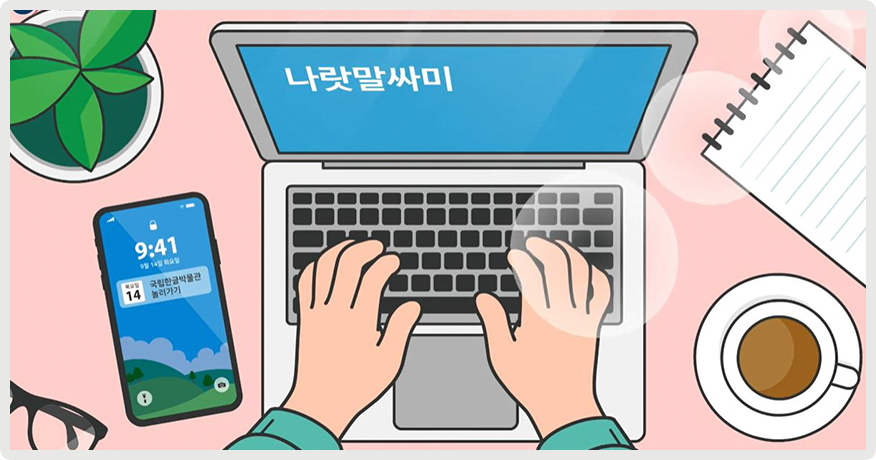 책상 위에 노트북과 노트, 화분, 커피, 핸드폰, 안경 등이 놓여있다. 한 손이 노트북 자판을 치고 있다. 노트북 화면에는 ‘나랏말싸미’가 적혀있다. 