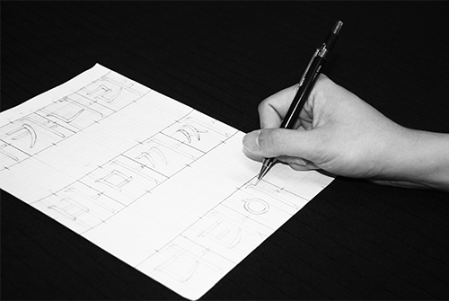 검은 배경에 흰 종이가 놓여있고 김정진 디자이너의 손이 연필을 쥔 채 글씨를 디자인하고 있다. 