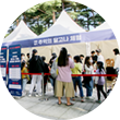 참여 행사 부스가 나란히 설치되어 있다. 그 앞에 수많은 사람이 줄을 서서 기다리고 있다. 사람들 주변에는 입간판이 놓여있다.
