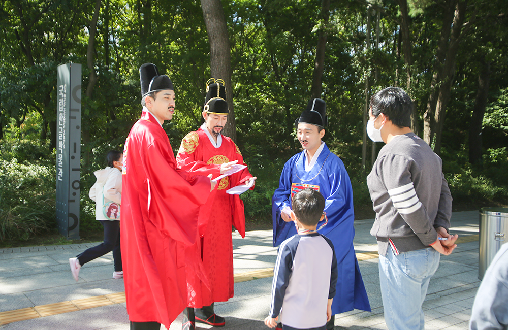 붉은색 곤룡포를 입은 세종대왕 역 배우가 손에 종이를 들고 앞에 선 아이에게 문제를 내고 있다. 주변으로는 파란색과 빨간색 조선시대 관료복을 관료 역 배우들이 미소짓고 있다. 아이의 뒤편에는 한 남성이 뒷짐을 진 채 배우들을 바라보고 있다.