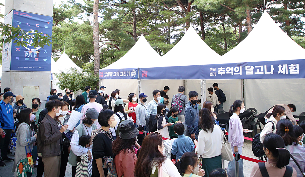 국립한글박물관으로 향하는 길목에 참여 행사 부스가 나란히 설치되어 있다. 길가에 설치된 기둥에는 ‘2022 한글주간’, ‘고마워 한글’이 적힌 포스터가 붙어있다. 부스 앞 길에는 사람들이 가득 차 있다.